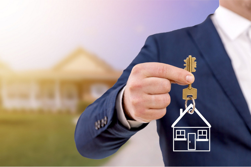 A person in a suit holds a key with a house-shaped keychain in front of a blurred background of a house.