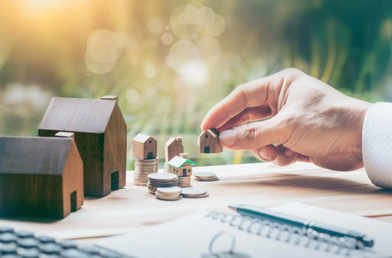 A hand holding a small wooden house near model homes and stacks of coins on a table.