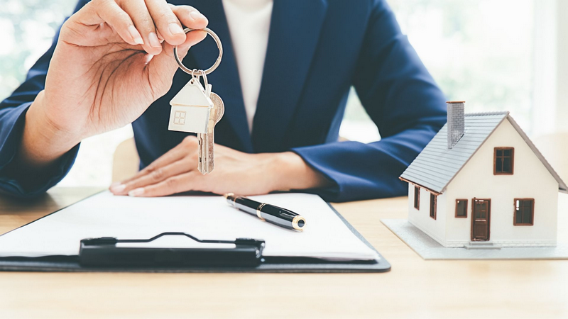 A person holding house keys over a clipboard with a small model house and a pen on a table.